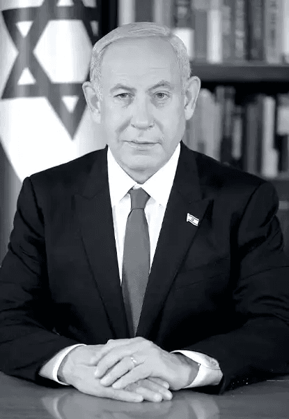 Fotografia oficial em preto e branco de Benjamin Netanyahu, primeiro-ministro de Israel, sentado à mesa com as mãos cruzadas. Ele veste terno escuro, camisa branca e gravata, com um pequeno distintivo da bandeira israelense no lapel. Ao fundo, à esquerda, parte da bandeira de Israel com a Estrela de Davi; atrás dele, estantes de livros. Sua expressão é séria, olhar direto para a câmera. A composição sugere autoridade, formalidade e poder institucional.