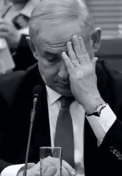 Fotografia em preto e branco de Benjamin Netanyahu, primeiro-ministro de Israel, sentado à mesa durante evento formal. Ele tem a mão direita levantada sobre o rosto, cobrindo os olhos e testa, como se estivesse cansado, pensativo ou sob pressão. Veste terno escuro, camisa branca e gravata. Usa relógio no pulso esquerdo e aliança no dedo anelar direito. À sua frente, há um microfone e um copo d'água. O fundo é desfocado, sugerindo ambiente institucional — possivelmente tribunal, parlamento ou sala de reuniões.