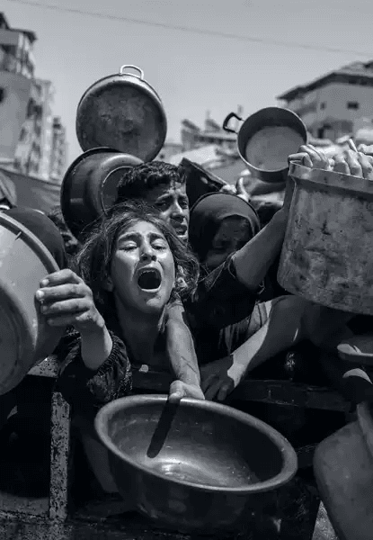 Fotografia em preto e branco mostrando grupo de crianças e jovens palestinos em estado de desespero, segurando panelas e bacias vazias acima da cabeça, como se pedissem comida ou água. No centro, uma menina grita com a boca aberta, olhos fechados, expressão de angústia extrema. Outros rostos ao redor mostram sofrimento, cansaço e urgência. Ao fundo, edifícios urbanos parcialmente destruídos indicam cenário de conflito. A imagem documenta a crise humanitária aguda em Gaza, onde civis enfrentam escassez severa de alimentos e recursos básicos.