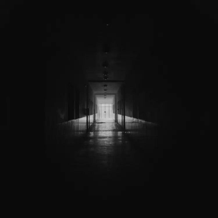 Corredor estreito e escuro em preto e branco. Uma luz intensa ao fundo cria um contraste dramático com as paredes laterais mergulhadas em sombras.