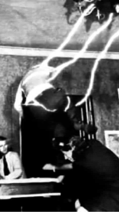 Fotografia P&B de uma pessoa levitando ou sendo arremessada sobre uma cama, com rastros de luz e movimento. Dois homens observam ao fundo em um ambiente doméstico.