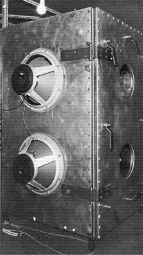Estrutura metálica industrial em P&B com dois grandes alto-falantes acoplados na frente, alavancas laterais e dobradiças pesadas. Objeto de aparência experimental.