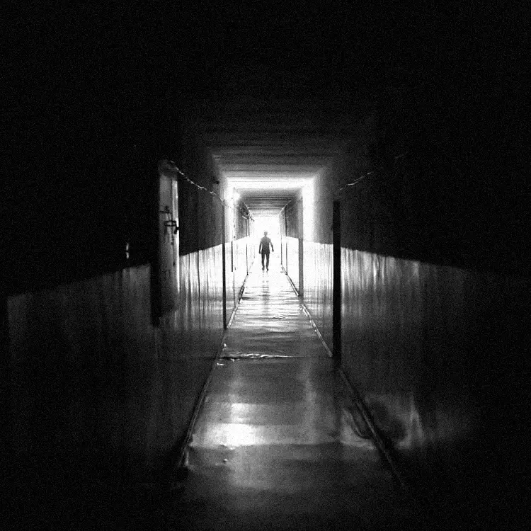 Corredor longo e granulado com luz ao fundo. Uma silhueta humana sombria observa imóvel de uma porta distante, criando tensão e medo.