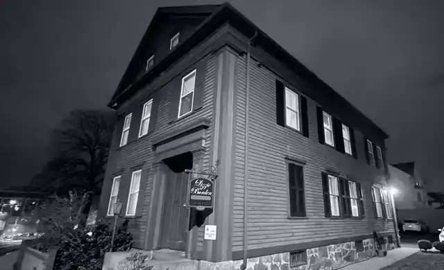 Vista noturna da residência em Massachusetts com a placa do museu visível. Atmosfera sombria ligada ao crime histórico.