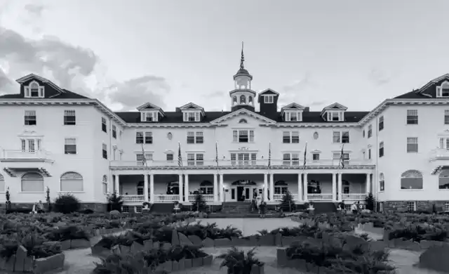 Fachada branca e simétrica do hotel que inspirou O Iluminado. Fotografia P&B nítida sob montanhas do Colorado.