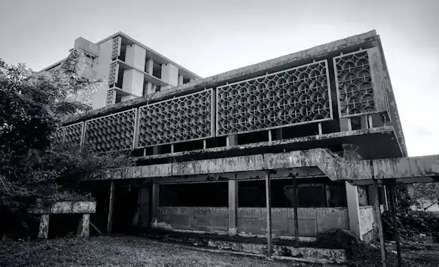 Edifício de arquitetura bruta abandonado e parcialmente tomado pela vegetação. Fotografia P&B de alto contraste e aura de mistério.