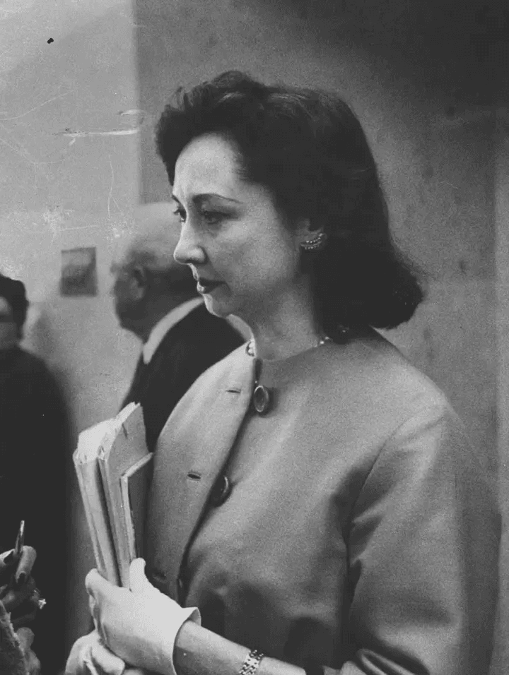 Mulher séria de perfil, com casaco e luvas brancas, segurando papéis. Fundo desfocado.