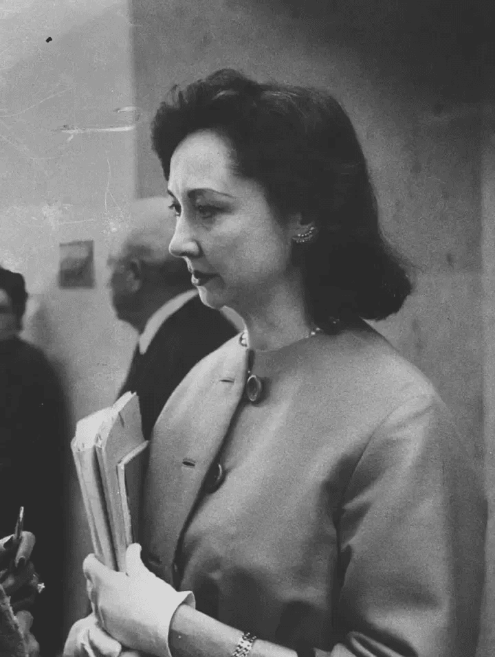 Mulher séria de perfil, com casaco e luvas brancas, segurando papéis. Fundo desfocado.