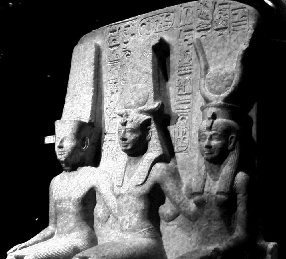 Tríade de estátuas egípcias esculpidas em pedra escura. Três figuras divinas sentadas em tronos, com coroas cerimoniais, transmitem uma aura de autoridade milenar e mistério.