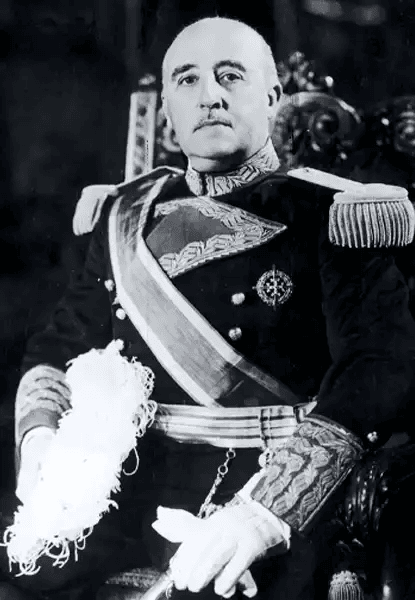Fotografia histórica em preto e branco do generalíssimo Francisco Franco, ditador da Espanha. Ele aparece sentado, de frente, com expressão severa e olhar fixo. Veste uniforme militar formal escuro, com ombreiras decoradas, faixa transversal bordada, luvas brancas e espada ao lado. No peito, medalhas e insígnias militares. O fundo é escuro e ornamentado, sugerindo trono ou cadeira real. Sua postura transmite autoridade absoluta.