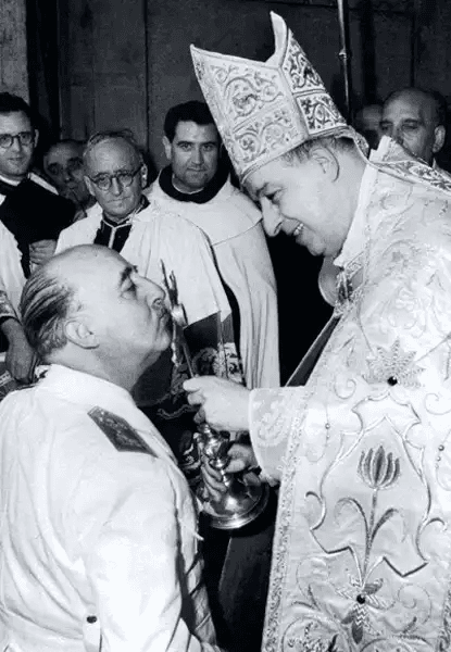 Fotografia histórica em preto e branco mostrando Francisco Franco, à esquerda, ajoelhado ou inclinado para beijar a mão de um bispo ou cardeal da Igreja Católica, vestido com ornamentos litúrgicos ricos: mitra bordada, casula decorada e segurando um cálice prateado. Ao fundo, outros sacerdotes e homens em trajes formais observam. Franco veste uniforme militar branco com distintivos no ombro. A expressão do clérigo é serena; a de Franco, reverente. A cena ocorre em ambiente interno, possivelmente catedral ou igreja.