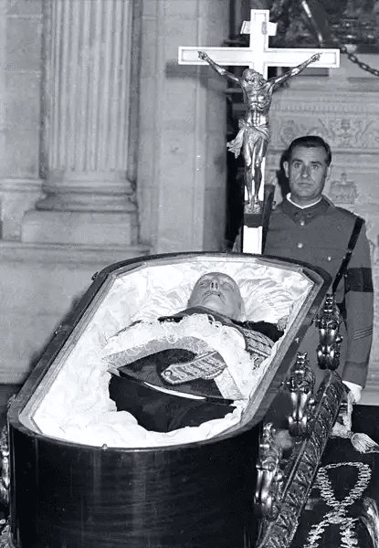 Fotografia histórica em preto e branco mostrando o corpo embalsamado de Francisco Franco dentro de um caixão aberto, vestido em uniforme militar formal com distintivos e luvas brancas. Ao lado direito, um soldado em uniforme cerimonial permanece em posição de guarda, olhando para frente com expressão séria. Ao fundo, uma grande cruz com estátua de Cristo crucificado domina a composição arquitetônica, sugerindo catedral ou mausoléu. O ambiente é solene, com colunas clássicas visíveis ao fundo.