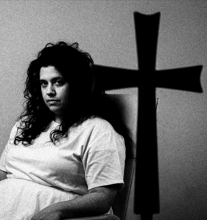 Fotografia em preto e branco de alto contraste. Uma mulher de olhar melancólico sentada ao lado da silhueta imponente de uma grande cruz negra que domina o cenário sombrio.