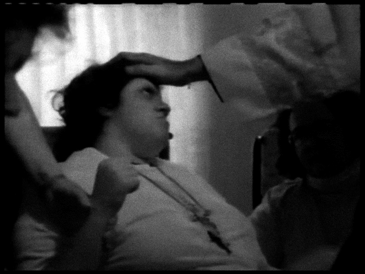Close-up granulado de uma mão pressionando a testa de uma mulher que parece estar em transe ou sofrimento. A estética de filme antigo reforça a tensão de um registro paranormal.