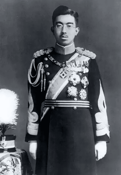 Fotografia histórica em preto e branco do Imperador Hirohito do Japão, em pé, de frente, com expressão séria e olhar direto. Ele veste uniforme militar formal escuro, ricamente decorado: ombreiras bordadas, cordões cerimoniais no peito, faixa transversal com insígnias, múltiplas medalhas e condecorações, e mangas com detalhes ornamentais. Usa óculos redondos e tem cabelo curto penteado para trás. Ao lado esquerdo, há uma mesa com objeto coberto por tecido bordado e pluma branca, possivelmente parte de ritual imperial. Fundo neutro e escuro, destacando sua figura central.
