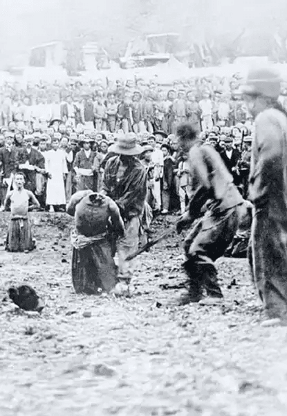 Fotografia histórica em preto e branco mostrando soldados japoneses executando civis chineses em Nanquim, China, em 1937–1938. No centro, um soldado ergue uma espada ou baioneta sobre uma vítima ajoelhada; outra vítima já está caída no chão. Ao fundo, uma grande multidão de civis observa passivamente, alguns com expressões de terror, outros com resignação. O cenário é ao ar livre, terreno irregular, possivelmente praça ou campo aberto. A imagem documenta crimes de guerra cometidos pelo Exército Imperial Japonês durante a ocupação de Nanquim.