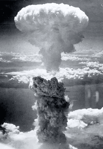 Fotografia histórica em preto e branco mostrando a nuvem em forma de cogumelo resultante da explosão de uma bomba atômica sobre o Japão em 1945. Uma coluna espessa de fumaça escura sobe verticalmente desde o solo, expandindo-se no alto em uma massa branca e volumosa contra um céu parcialmente nublado. Não há cidades visíveis abaixo — apenas a imensidão da destruição atmosférica. A imagem foi tirada de um avião, capturando o momento exato da detonação nuclear.