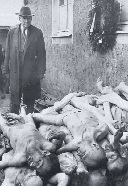 Fotografia histórica em preto e branco mostrando um homem civilizado, vestido com casaco longo e chapéu, parado ao lado de uma pilha de corpos esqueléticos de vítimas de campo de concentração. Os cadáveres estão empilhados no chão, nus ou parcialmente cobertos, com ossos visíveis e expressões de sofrimento final. Ao fundo, há uma parede de edifício simples e uma coroa fúnebre pendurada na janela. A cena transmite o choque e a gravidade do genocídio nazista.