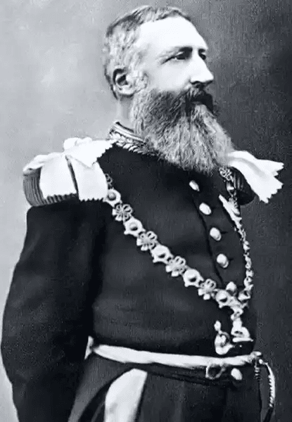 Fotografia histórica em preto e branco do rei Leopoldo II da Bélgica, retratado de perfil. Ele tem barba longa e grisalha, cabelos curtos penteados para trás, e veste uniforme militar formal com ombreiras brancas, botões metálicos e uma corrente cerimonial ornamental cruzando o peito. Sua expressão é séria e distante, olhando para fora do quadro.