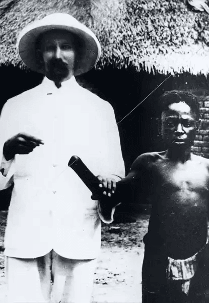 Fotografia histórica em preto e branco mostrando um homem europeu, vestido com terno branco e chapéu de palha, segurando a mão amputada de um homem congolês descalço e sem camisa, que está ao seu lado. O homem congolês olha diretamente para a câmera; sua expressão é séria e contida. Ao fundo, há uma cabana de telhado de palha. A imagem documenta a prática de mutilação como punição ou prova de trabalho forçado durante o Estado Livre do Congo.