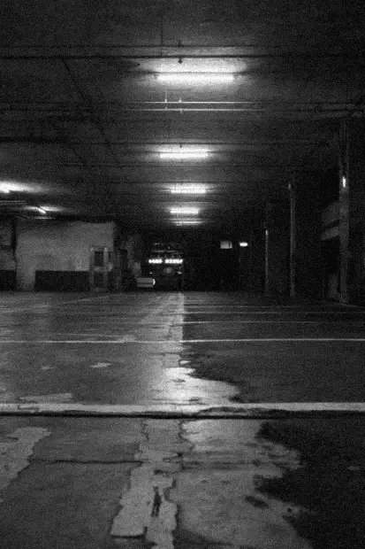Ambiente subterrâneo escuro e úmido. Luzes fluorescentes no teto refletem em poças no chão de concreto, sugerindo um local abandonado.