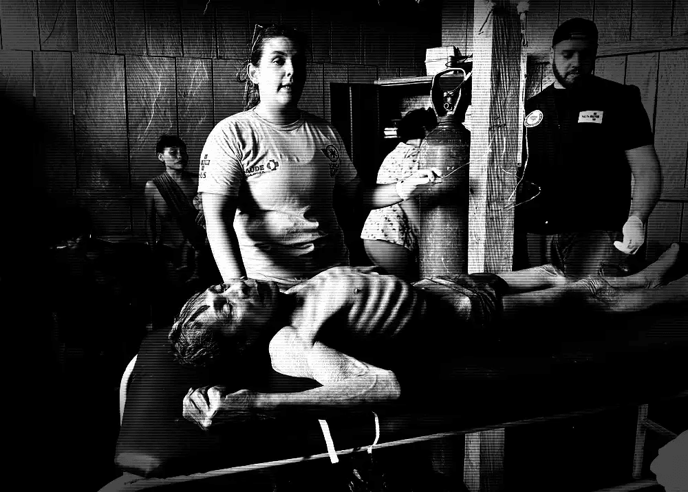 Cena de alto contraste em um posto de saúde. Profissionais médicos atendem uma pessoa indígena extremamente debilitada e desnutrida, deitada em uma maca sob iluminação dramática e sombria.