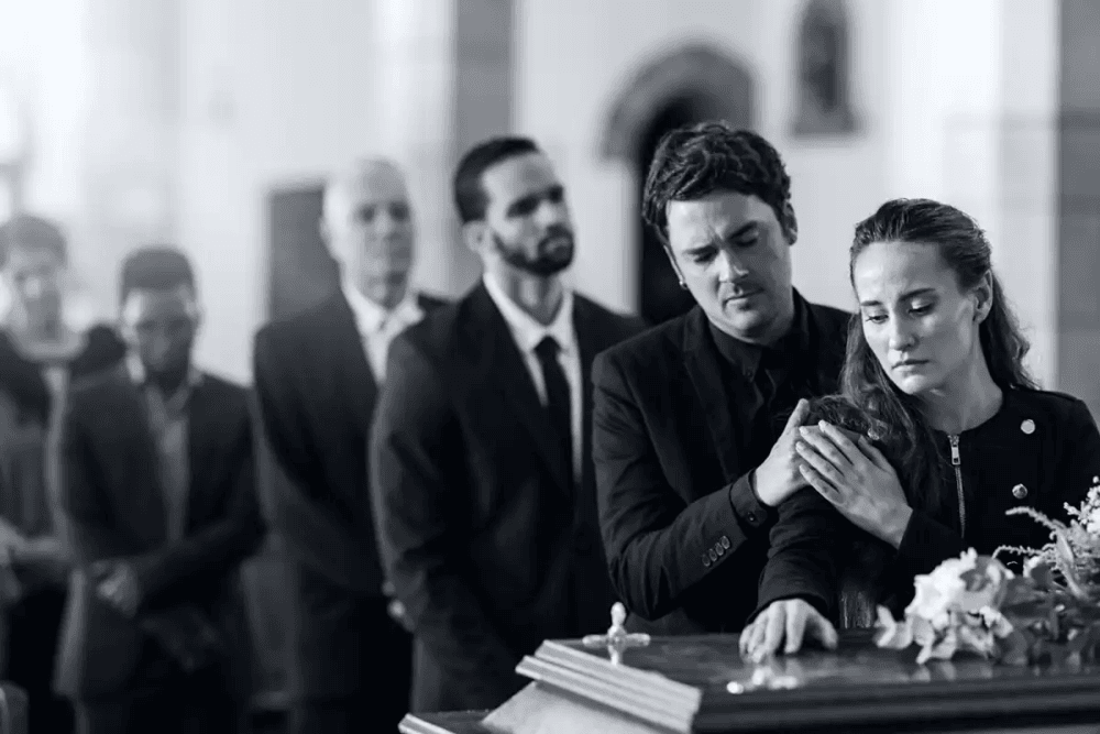 otografia P&B de funeral moderno. Um homem consola uma mulher em prantos que toca o caixão. A iluminação dramática destaca a dor e o luto em um ambiente eclesiástico.