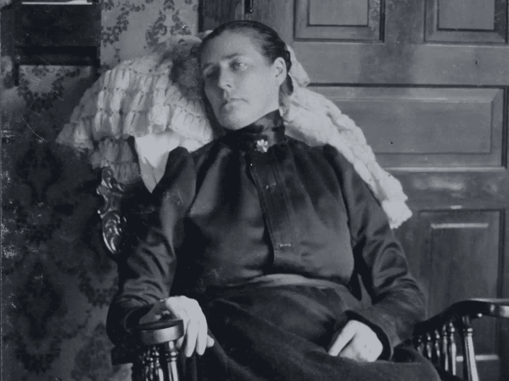 Fotografia vitoriana pós-morte: uma mulher pálida de negro sentada em uma poltrona. O olhar fixo e a rigidez capturam o costume sombrio de retratar os mortos como vivos.