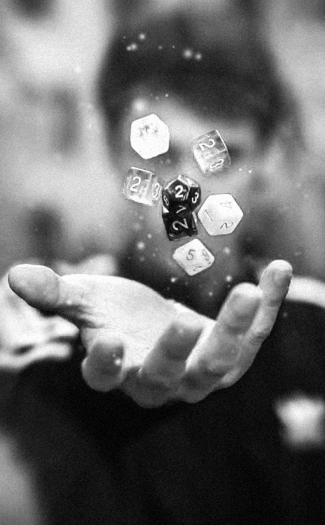 Close-up em preto e branco de uma mão capturando diversos dados multifacetados que flutuam no ar. A granulação da foto confere um tom enigmático sobre sorte, destino e jogos de RPG.
