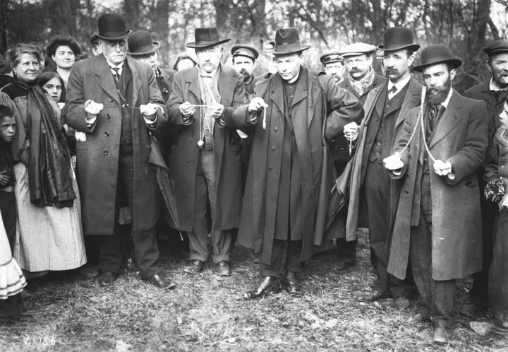 Homens de chapéu seguram pêndulos em bosque. Mulheres e crianças observam. Cena histórica sugere radiestesia.