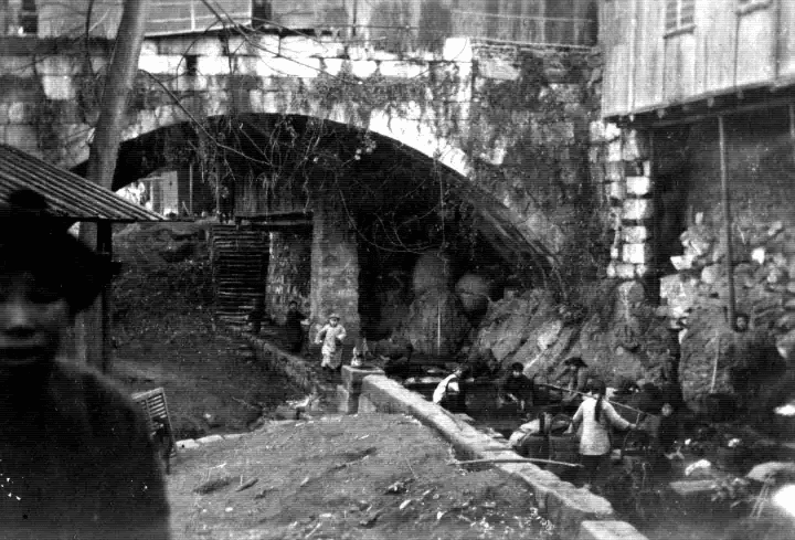 Cena P&B sob o arco de uma ponte de pedra antiga. Pessoas pequenas caminham ao fundo, enquanto um rosto desfocado em primeiro plano cria uma sensação de aparição.