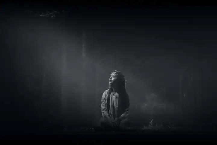 A Aparição: Jovem sentada em floresta escura e enevoada, sob um feixe de luz vertical. Fotografia P&B com atmosfera sobrenatural, melancólica e espiritual.