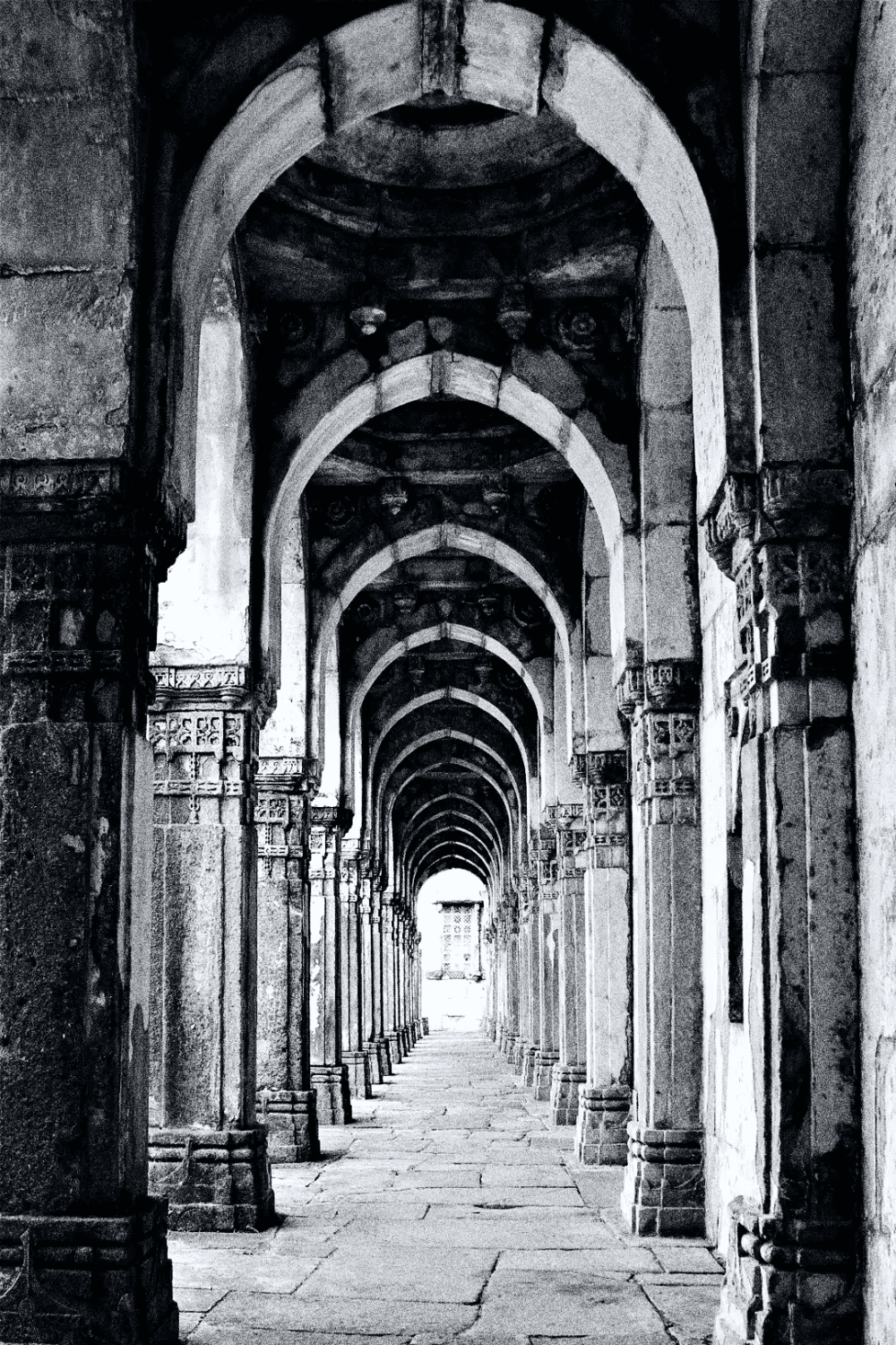 Corredor de arcos ancestrais em alto contraste P&B. Pilares entalhados formam uma perspectiva profunda e sombria rumo a uma luz intensa. Atmosfera de mistério, silêncio e arquitetura antiga.