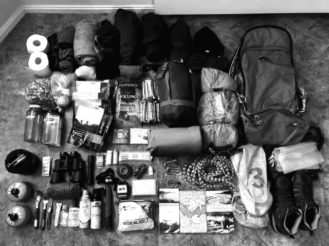 Equipamentos de sobrevivência organizados no chão: mochila, mantimentos, binóculos, kit médico, cordas e botas. Preparação minuciosa para uma jornada rumo ao desconhecido.