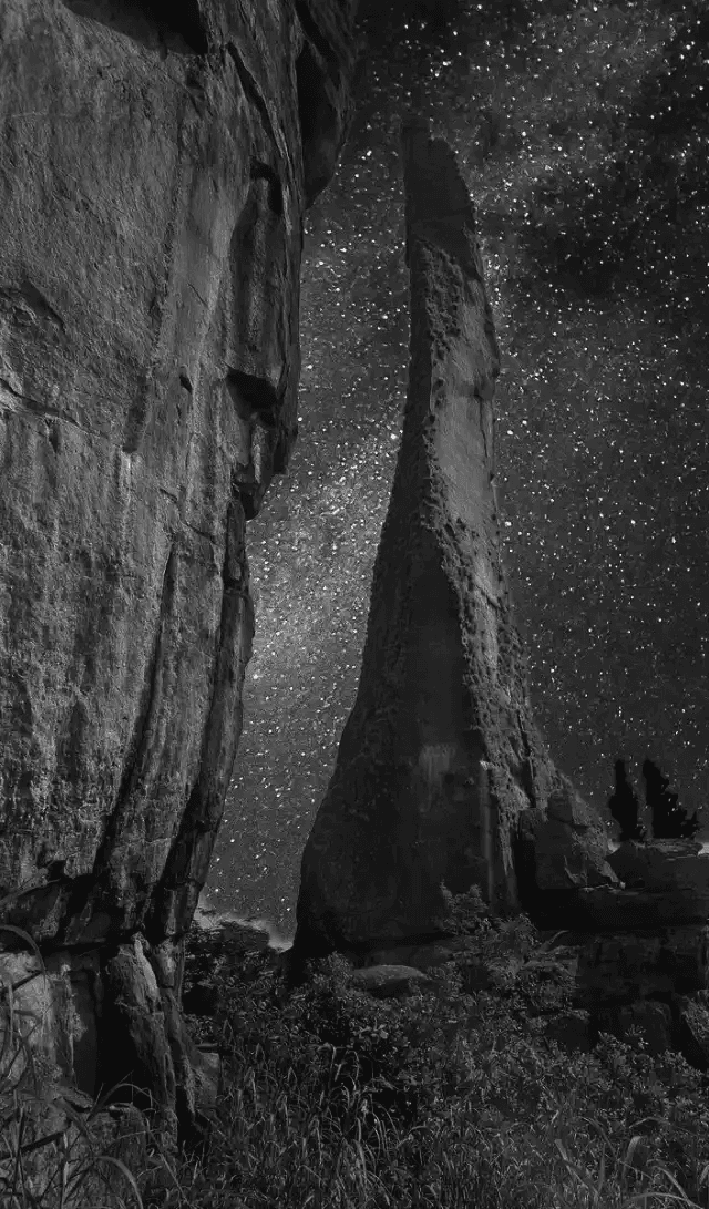 Formação rochosa vertical e pontiaguda elevando-se em direção a um céu noturno densamente estrelado. A cena transmite uma sensação de isolamento cósmico e antiguidade.