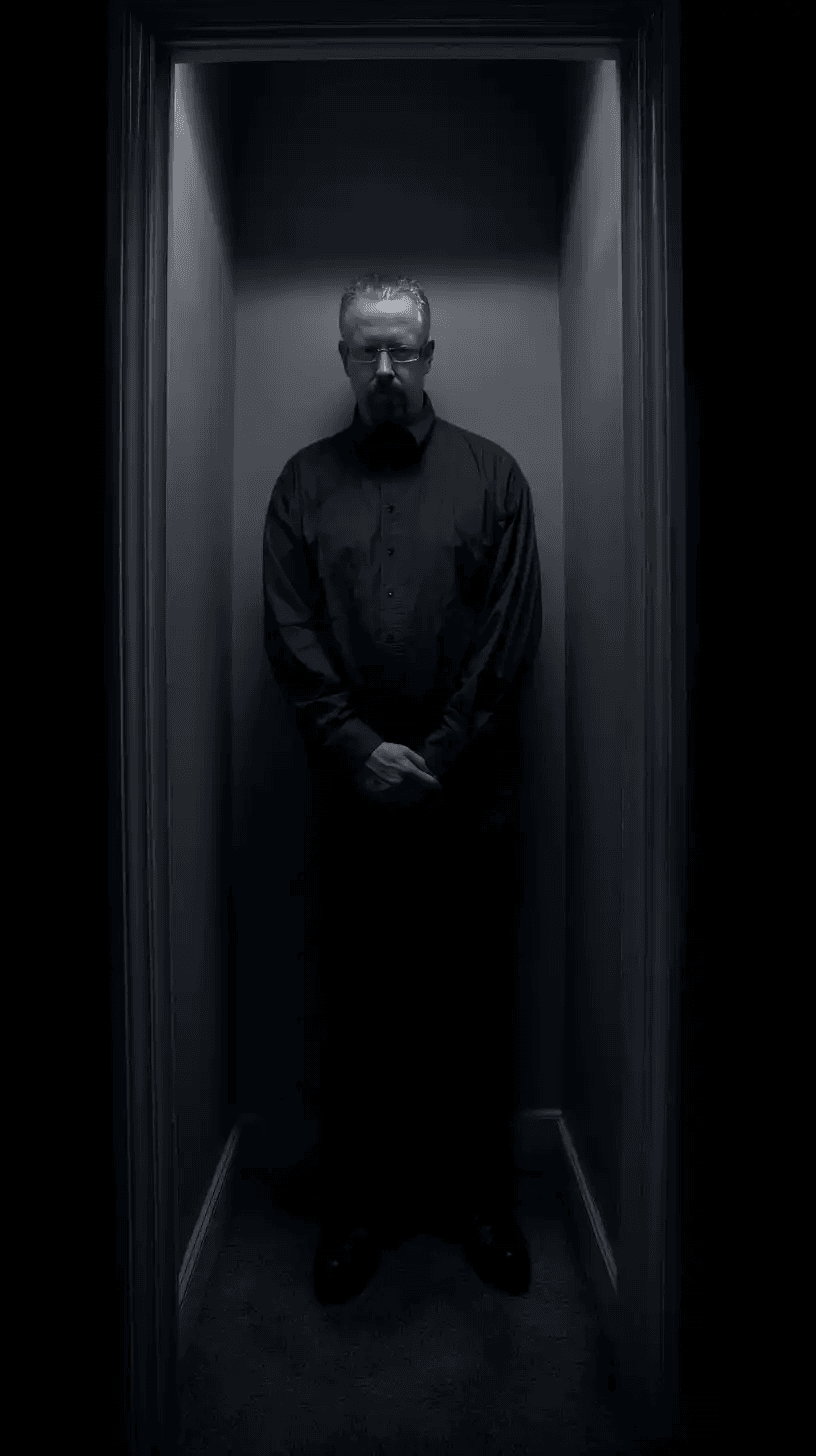 Homem de óculos e camisa escura posicionado em um nicho estreito sob luz zenital dramática. Sua expressão é severa e o ambiente é cercado por sombras profundas, evocando mistério.