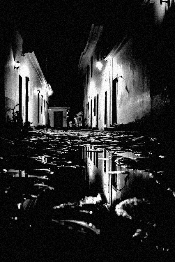 Uma rua de paralelepípedos escura com reflexos de poças, ladeada por edifícios baixos iluminados.