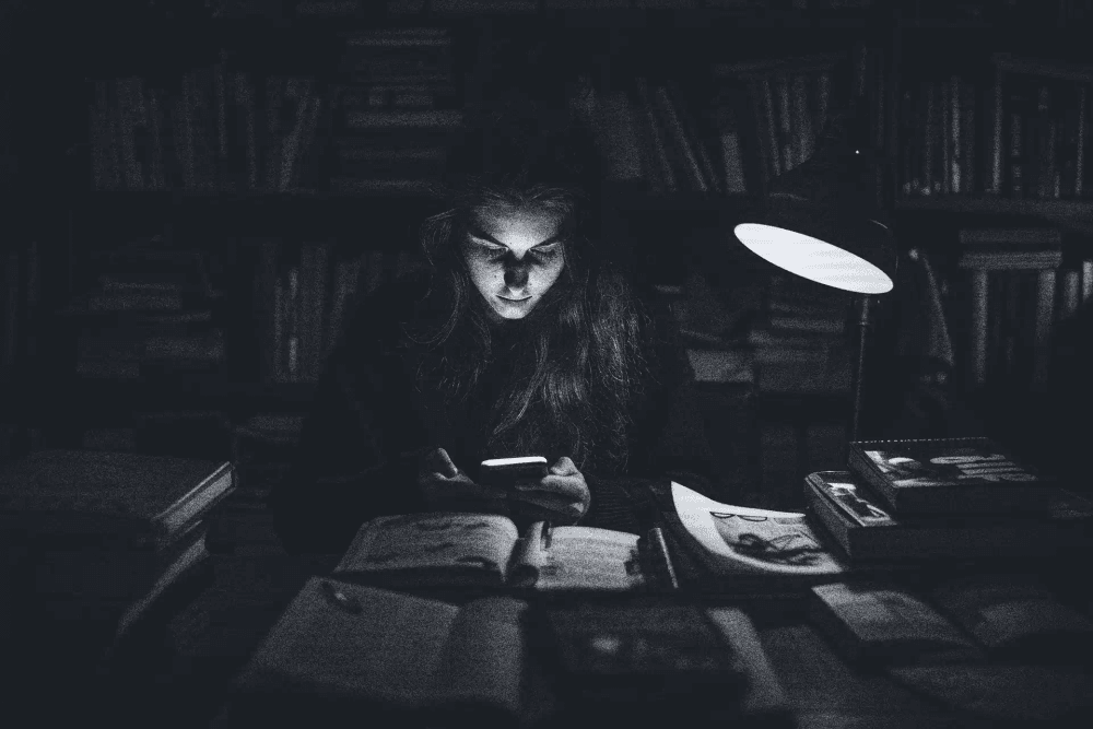 Jovem em uma biblioteca escura, iluminada apenas pela luz de um celular e uma luminária de mesa. O clima é de mistério e busca por conhecimento oculto entre livros.